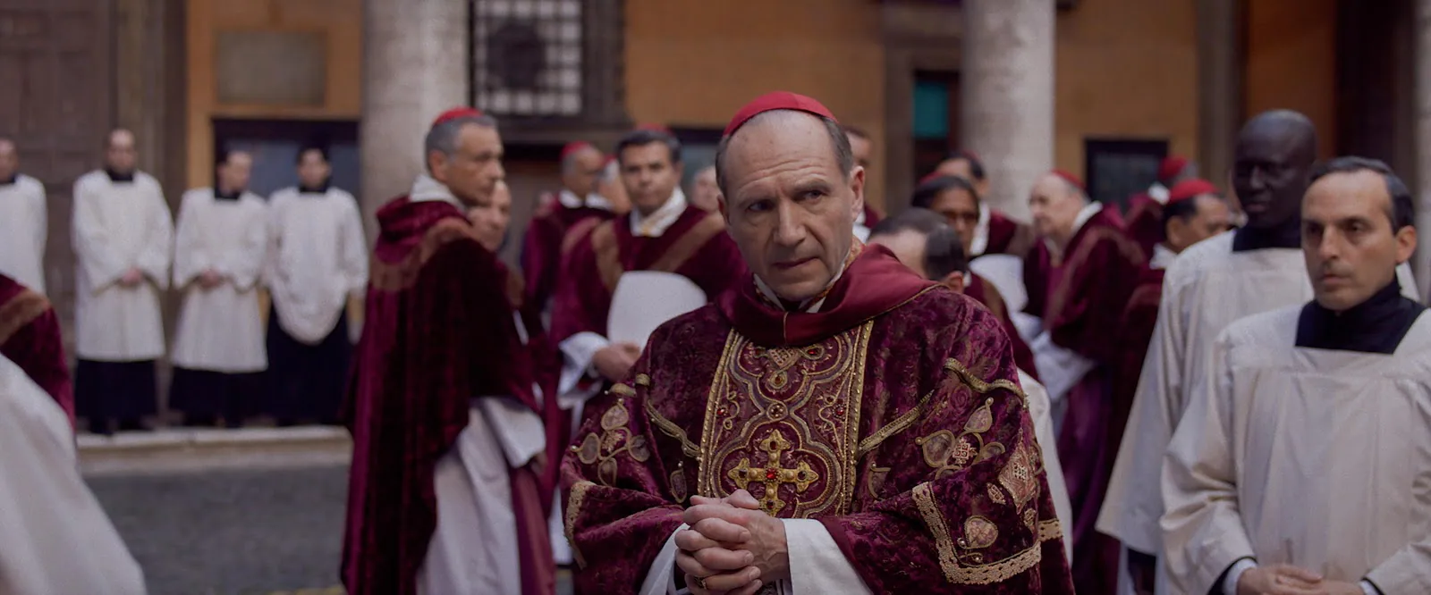 Ralph Fiennes in clerical black stands alone in a cavernous vaulted stone hall looking upward