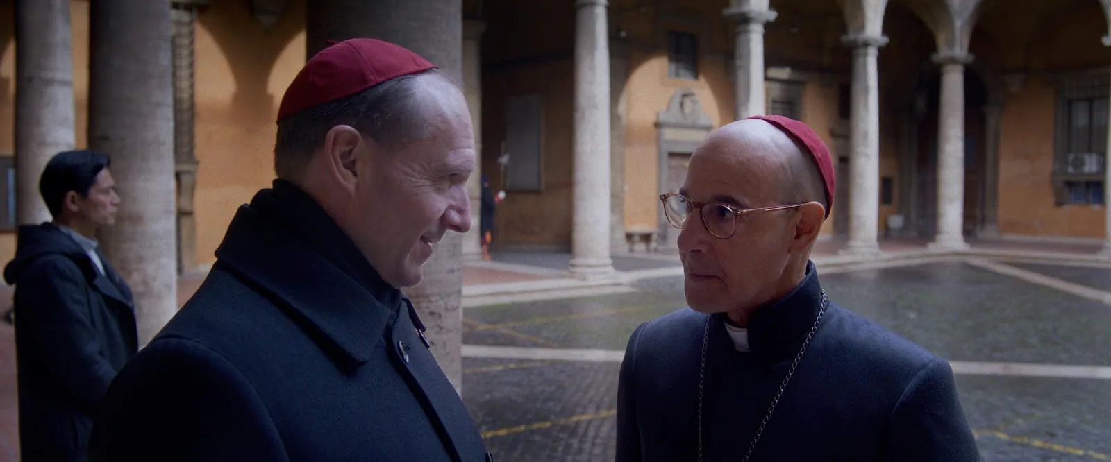 Ralph Fiennes and Stanley Tucci stand face to face in cardinal vestments in a pale formal corridor